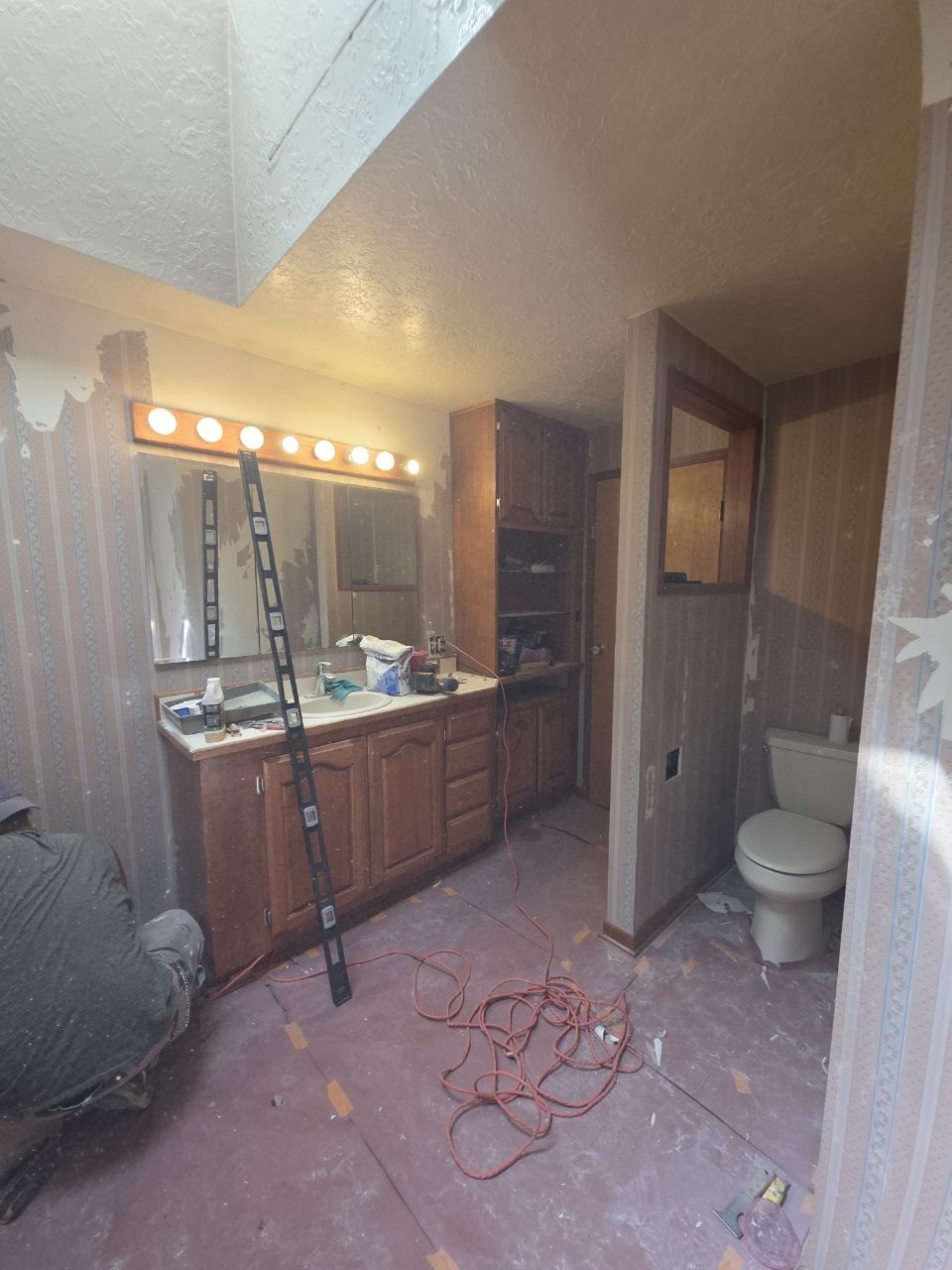 Primary bathroom vanity before remodel — dated wallpaper, cabinets, and lighting